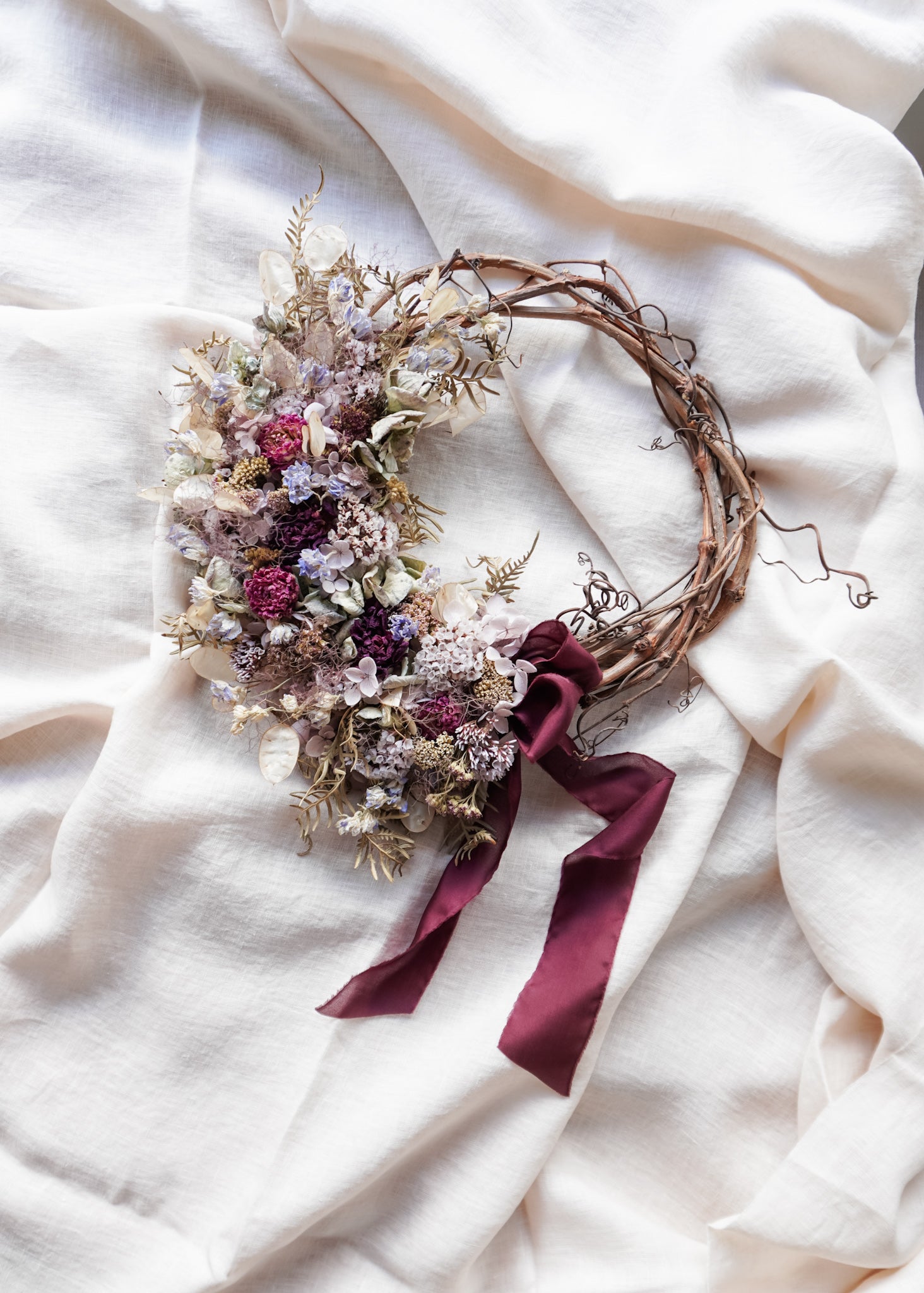 Dried flower wreath in blue violet tones on a grapevine base with burgundy ribbon.