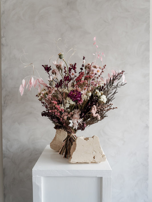 Bouquet of dried flowers in pink and burgundy tones on a white plinth against a grey background.