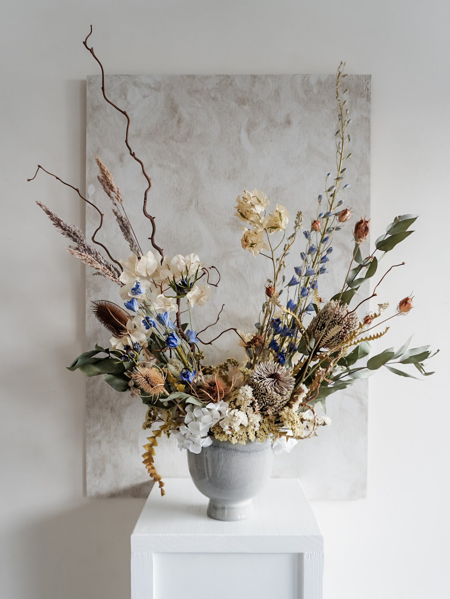 Dried flower arrangement in ceramic vase with natural toned florals accented with blue delphinium – full view.