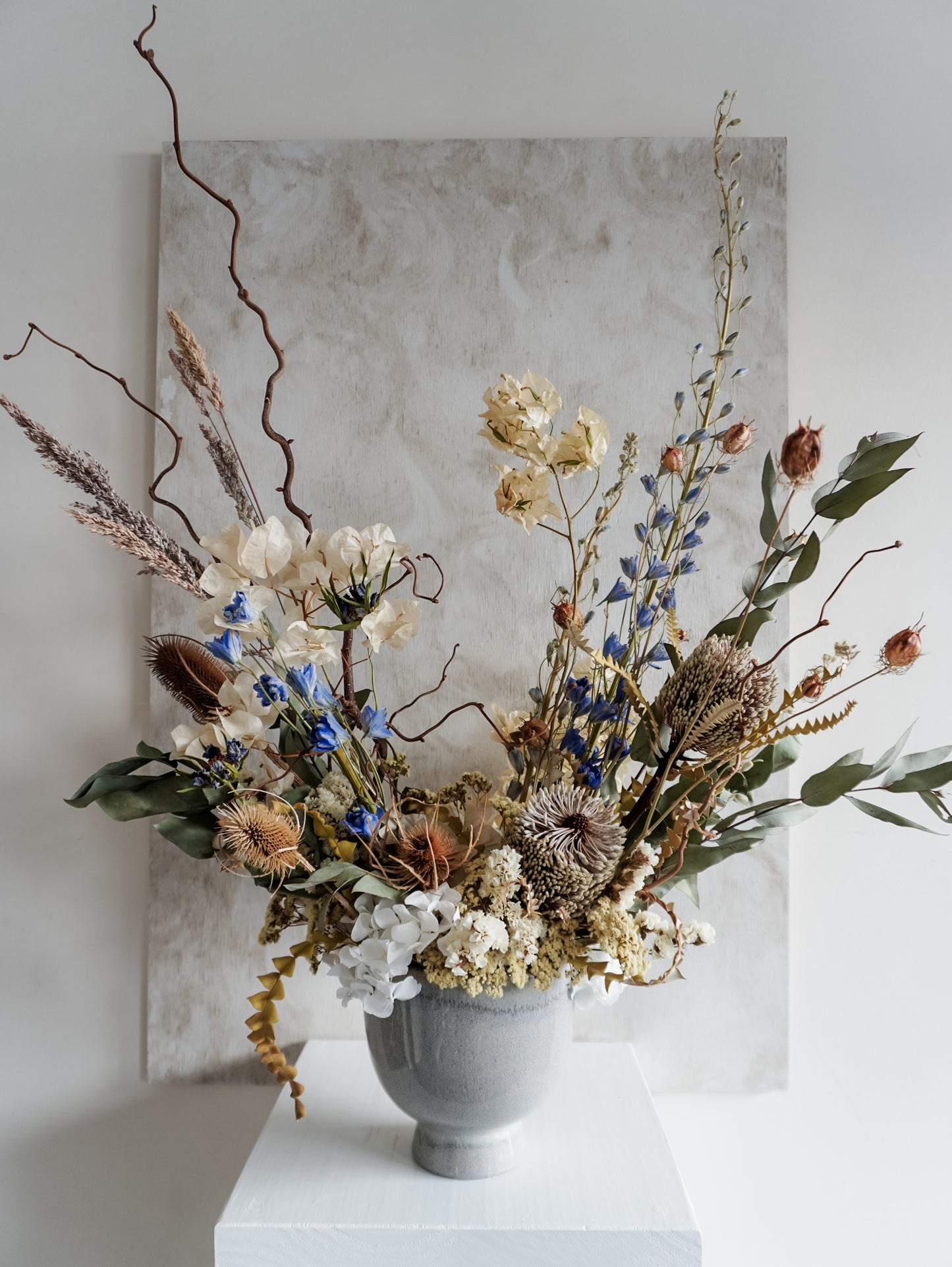 Dried flower arrangement in ceramic vase with natural toned florals accented with blue delphinium – close full view.