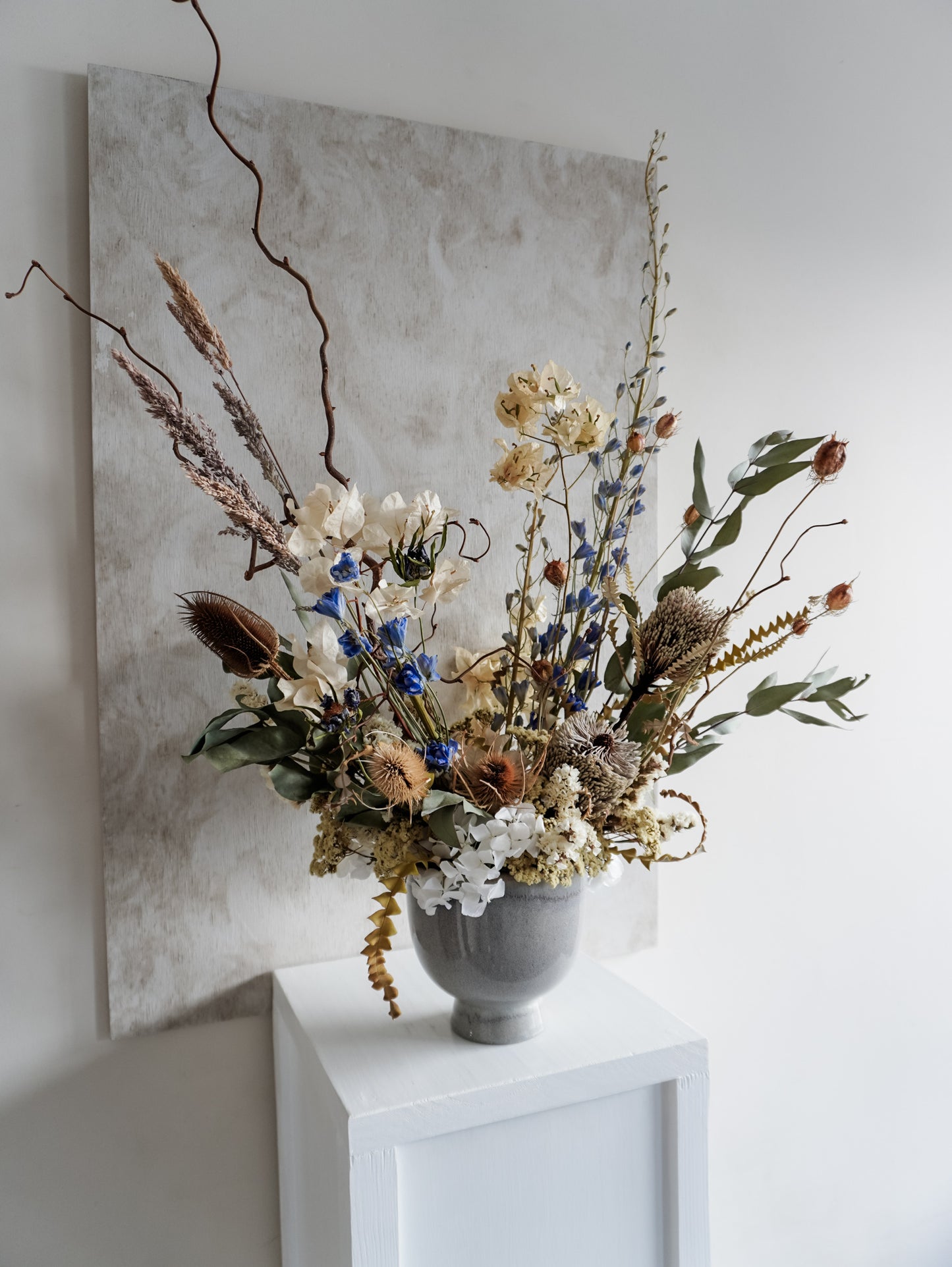 Dried flower arrangement in ceramic vase with natural toned florals accented with blue delphinium – left side view.