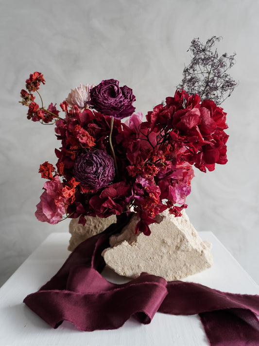 Bridesmaid bouquet of dried red and purple flowers on a white plinth with a gray background.