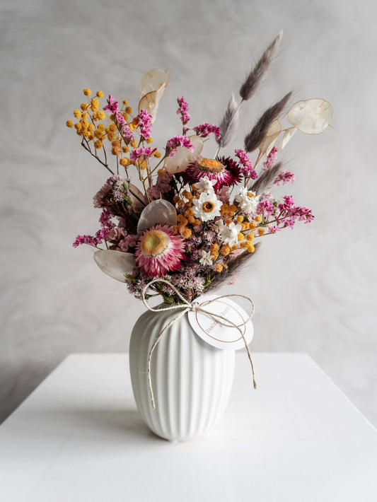 Dried flower arrangement in bright colours displayed in a white bud vase and positioned on a white plinth against a gray background. Close view.