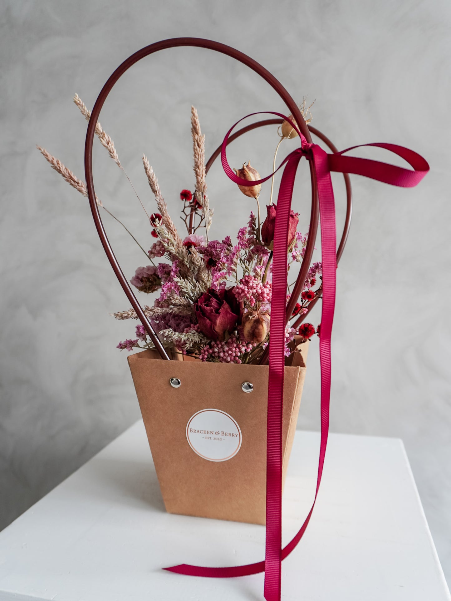 Dried floral arrangement in a kraft posy bag with a red ribbon on a white surface.