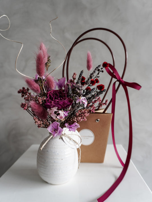 Dried flower arrangement in a white vase with a brown gift bag and pink ribbon on a light gray background