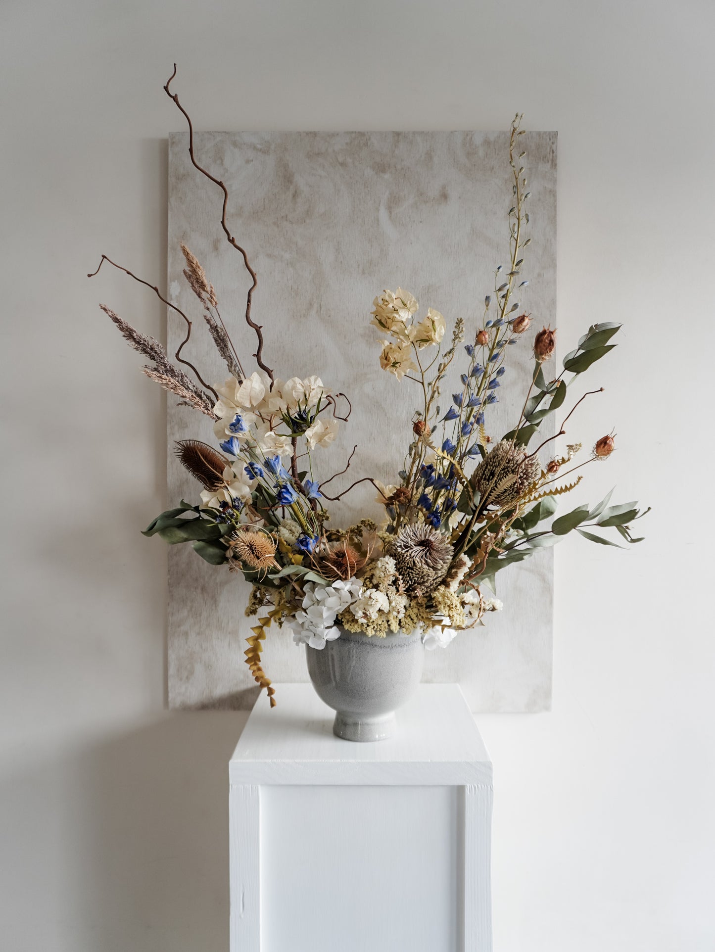 Dried flower arrangement in ceramic vase with natural toned florals accented with blue Delphinium – full view.