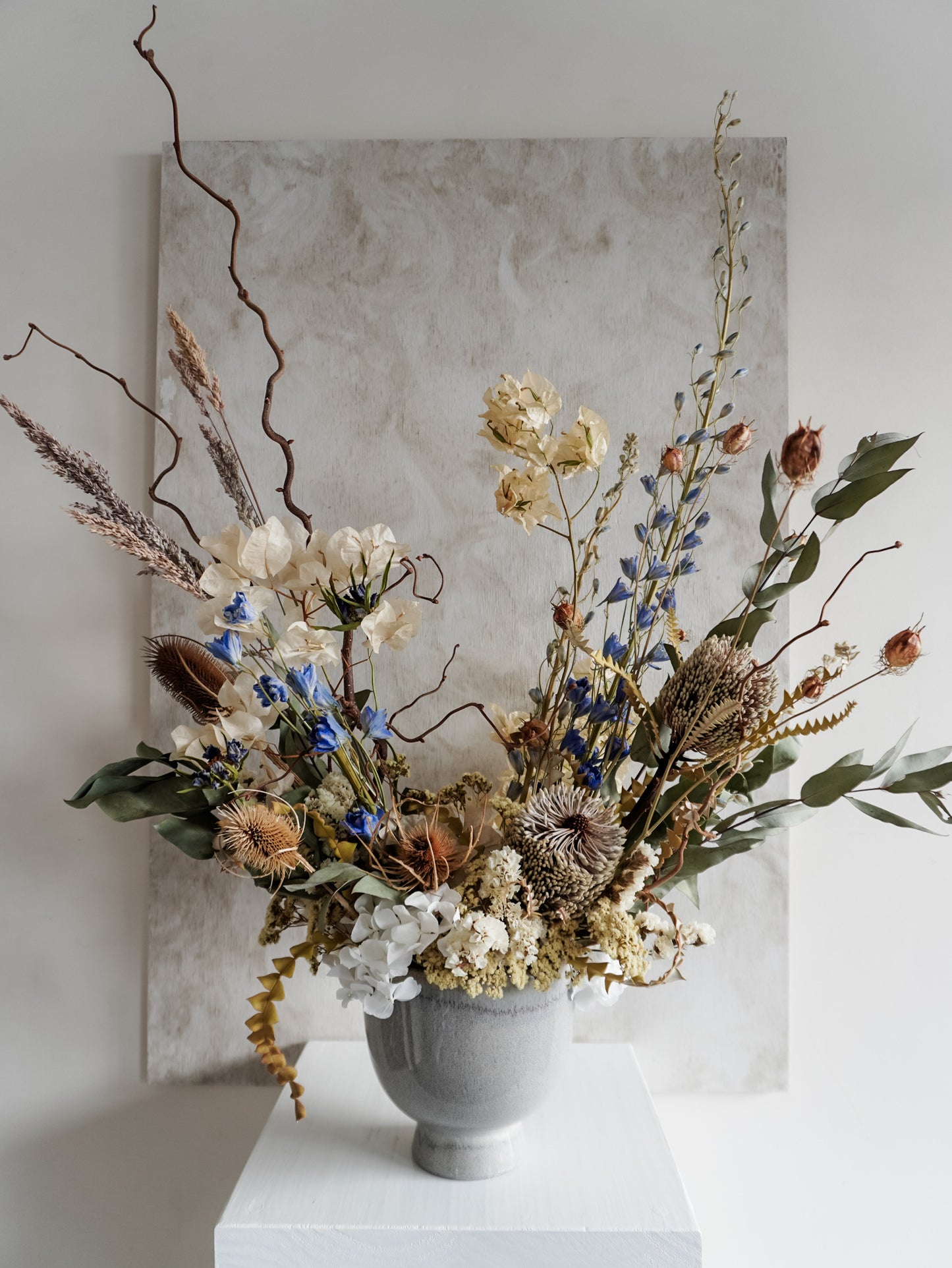 Dried flower arrangement in ceramic vase with natural toned florals accented with blue Delphinium – close view.