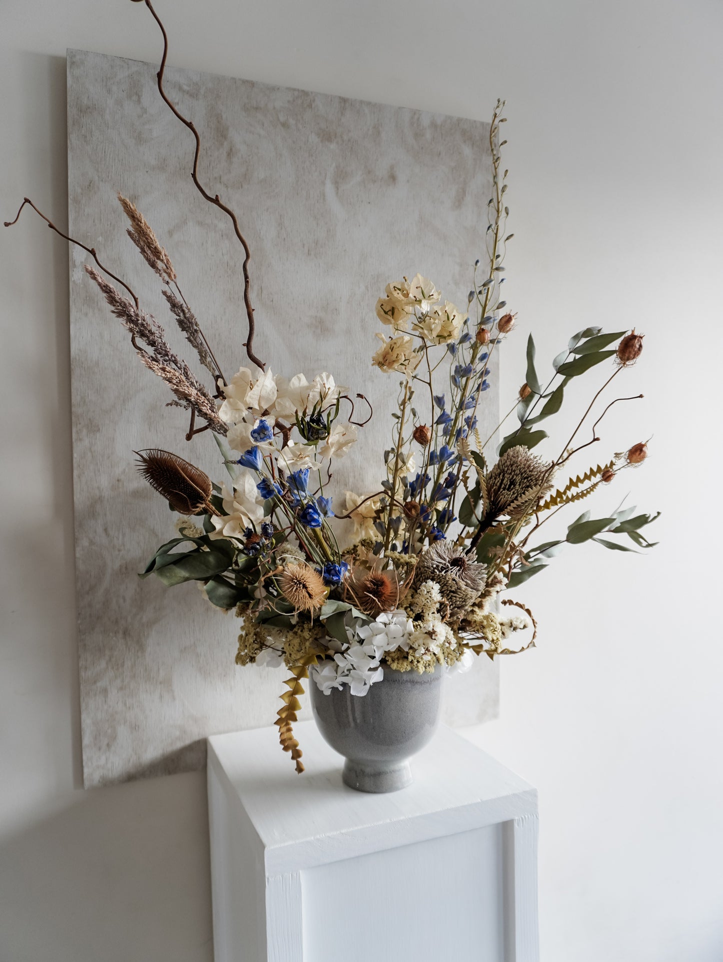 Dried flower arrangement in ceramic vase with natural toned florals accented with Blue delphinium – left angle view.