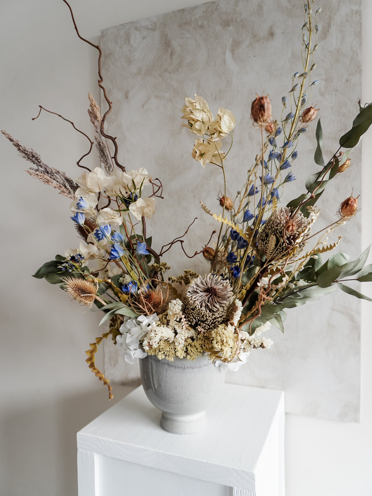 Dried flower arrangement in ceramic vase with natural toned florals accented with blue Delphinium – right angle view.