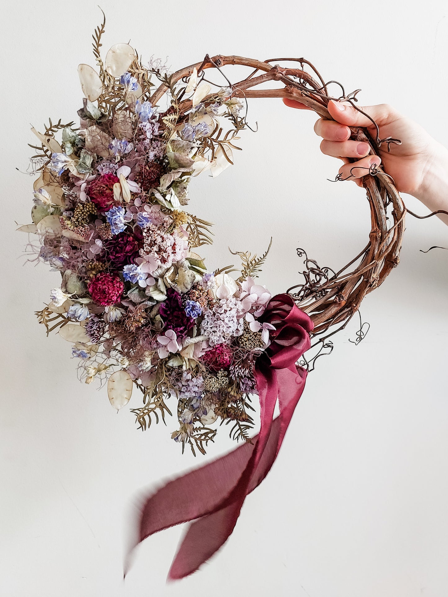 Dried flower wreath in rich blue violet tones on a grapevine base with a burgundy silk ribbon.