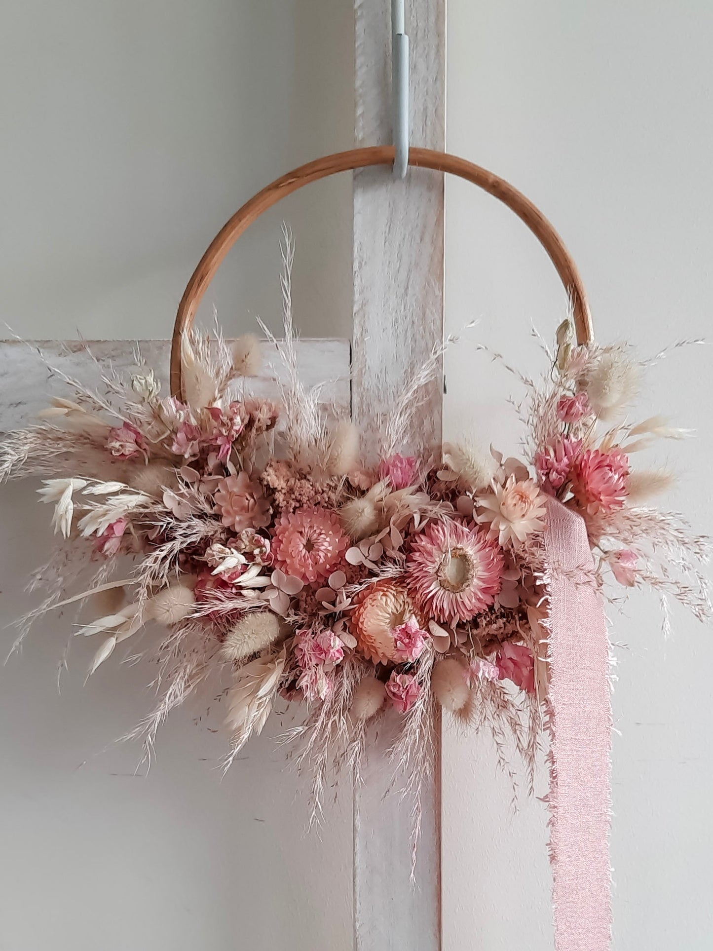 A rustic dried flower wreath featuring soft pink and cream dried flowers and foliage on a rattan hoop base, hanging on a white wooden ladder.