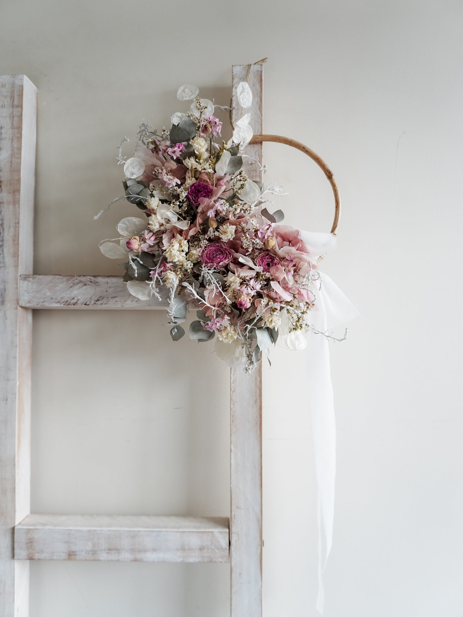 Dried flower wreath in soft pastel tones on a rattan hoop base with white ribbon - distant full view.