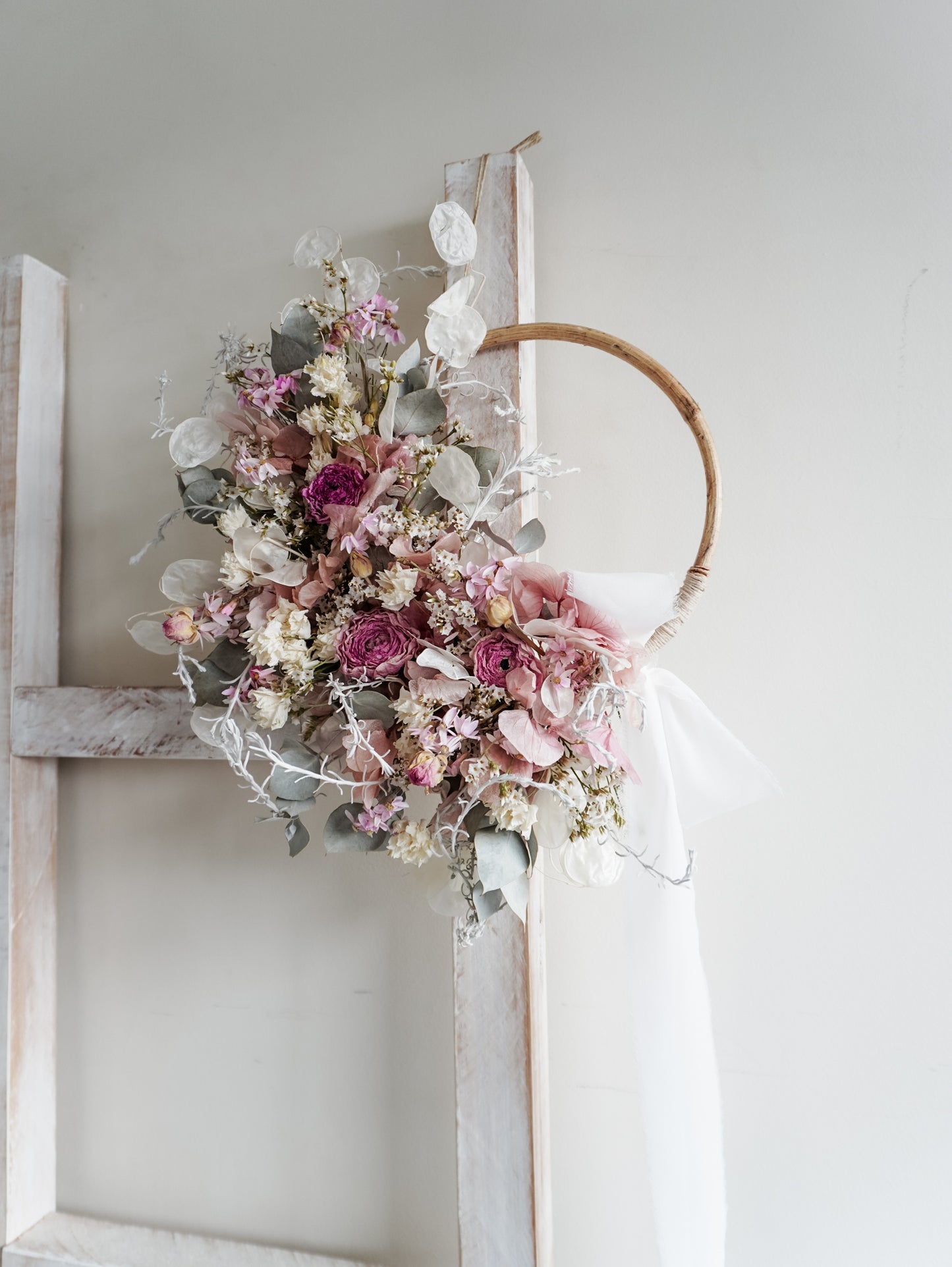 Dried flower wreath in soft pastel tones on a rattan hoop base with white ribbon - side view.