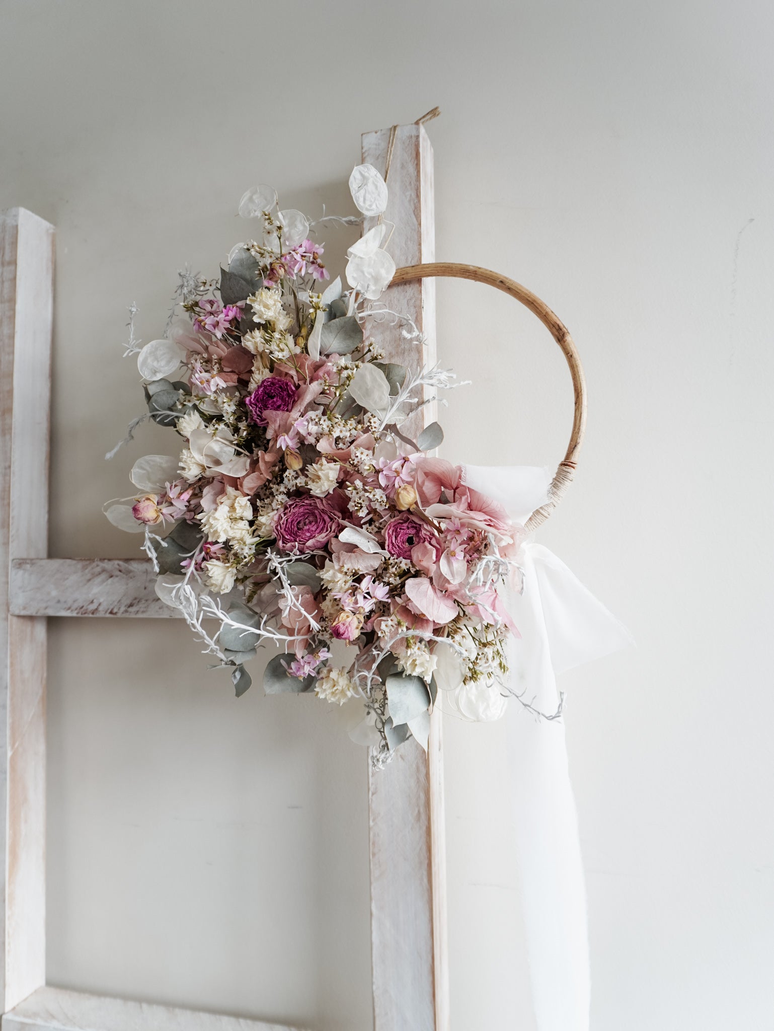 Dried flower wreath in soft pastel tones on a rattan hoop base with white ribbon - side view.