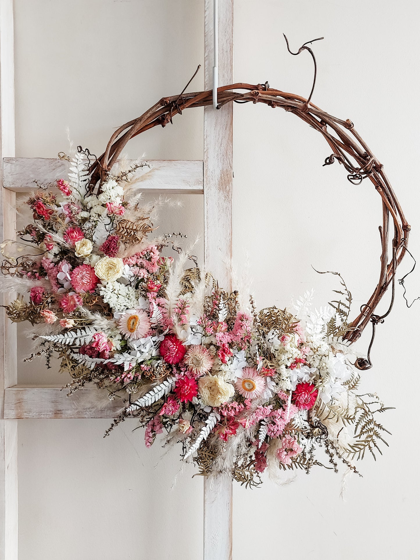 A rustic dried flower wreath featuring soft pink, white, and spring green dried flowers and foliage on a grapevine base, hanging on a white wooden ladder.