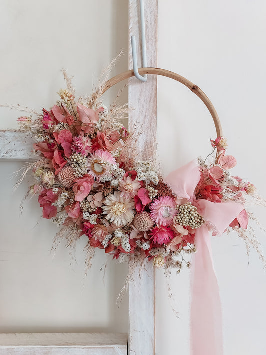 A rustic dried flower wreath featuring soft pink and cream dried flowers and foliage on a rattan hoop base, hanging on a white wooden ladder.