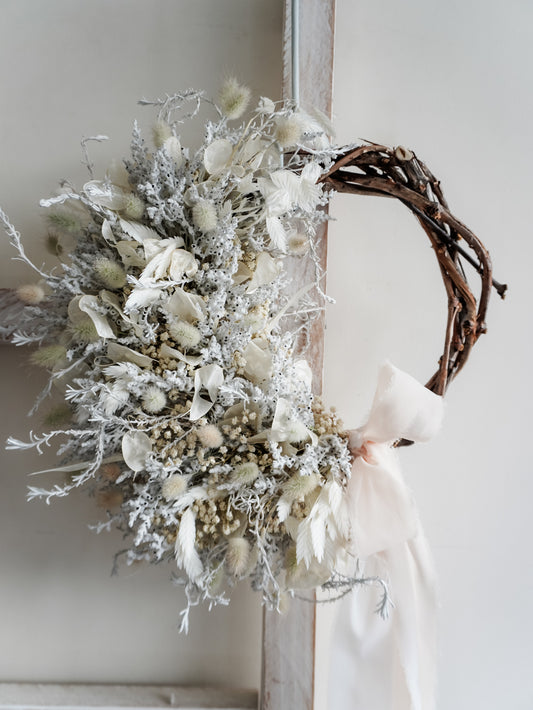 A rustic dried flower wreath featuring white, cream, and silver dried flowers and foliage on a grapevine base, hanging on a white wooden ladder.