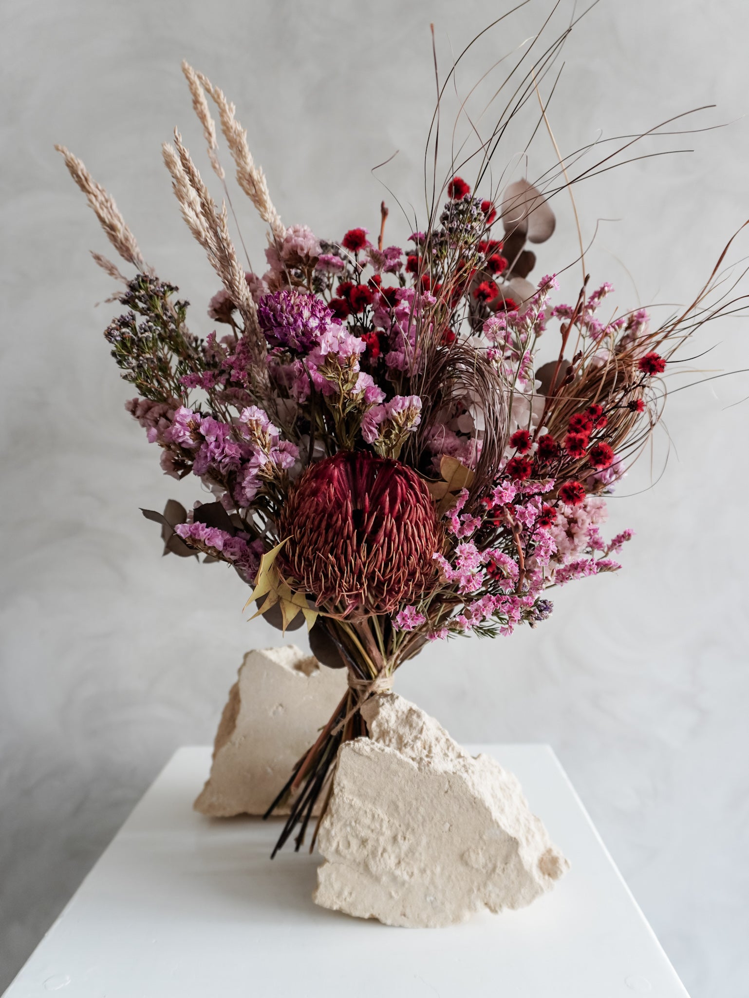 Bouquet of dried flowers in red and purple tones on a white plinth with a light gray background.
