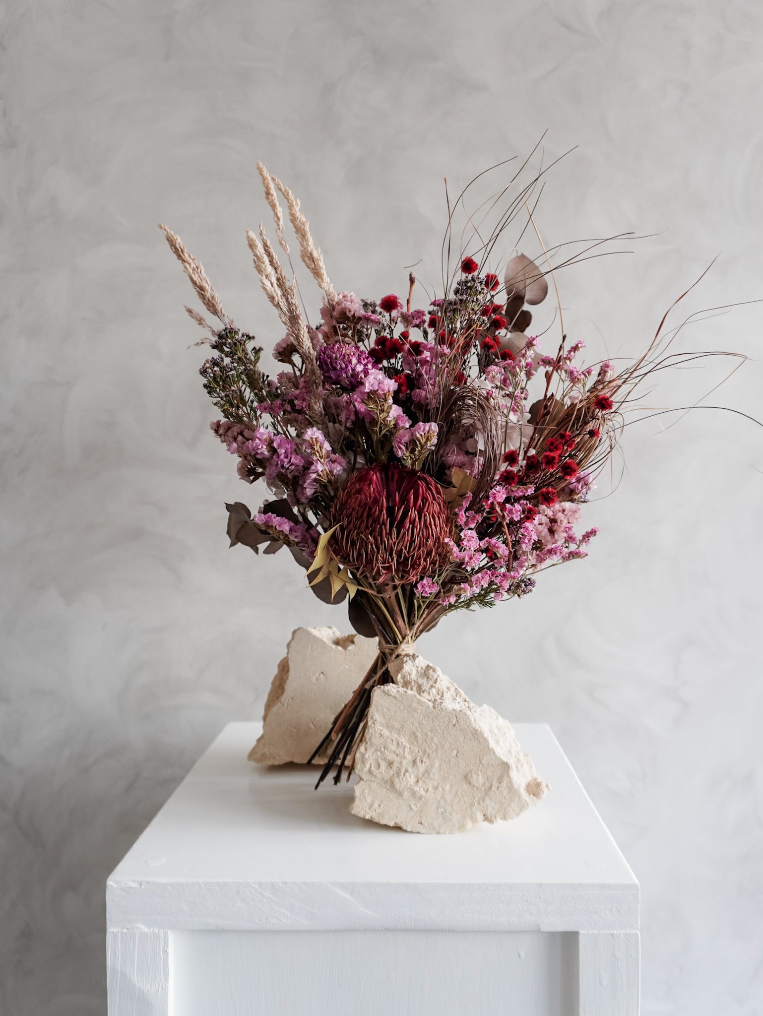 Bouquet of dried flowers in red and purple tones on a white plinth with a light gray background.