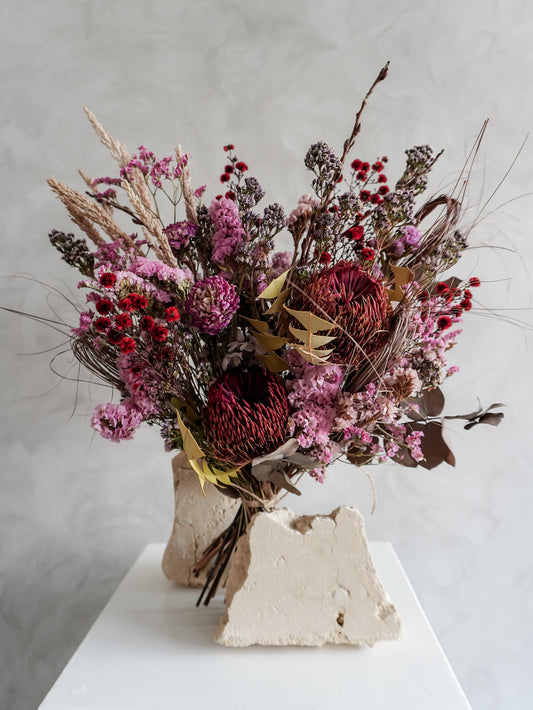 Bouquet of dried flowers in red and purple tones on a white surface with a light gray background.