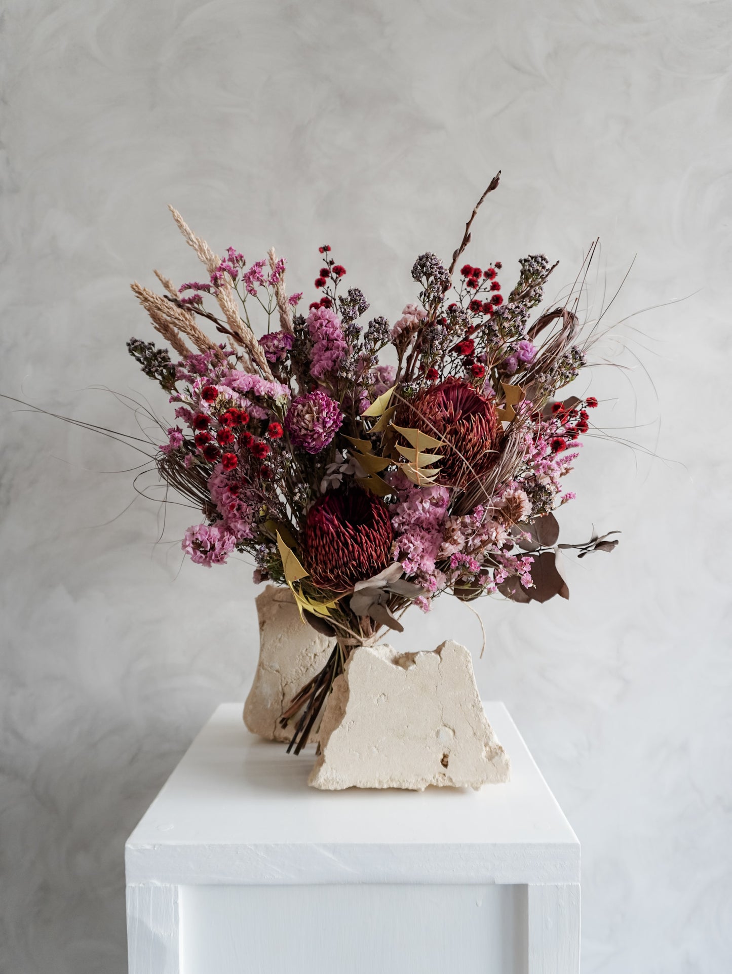 Bouquet of dried flowers in red and purple tones on a white plinth with a light gray background.