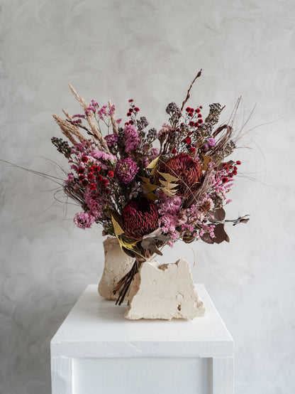 Bouquet of dried flowers in red and purple tones on a white plinth with a light gray background.