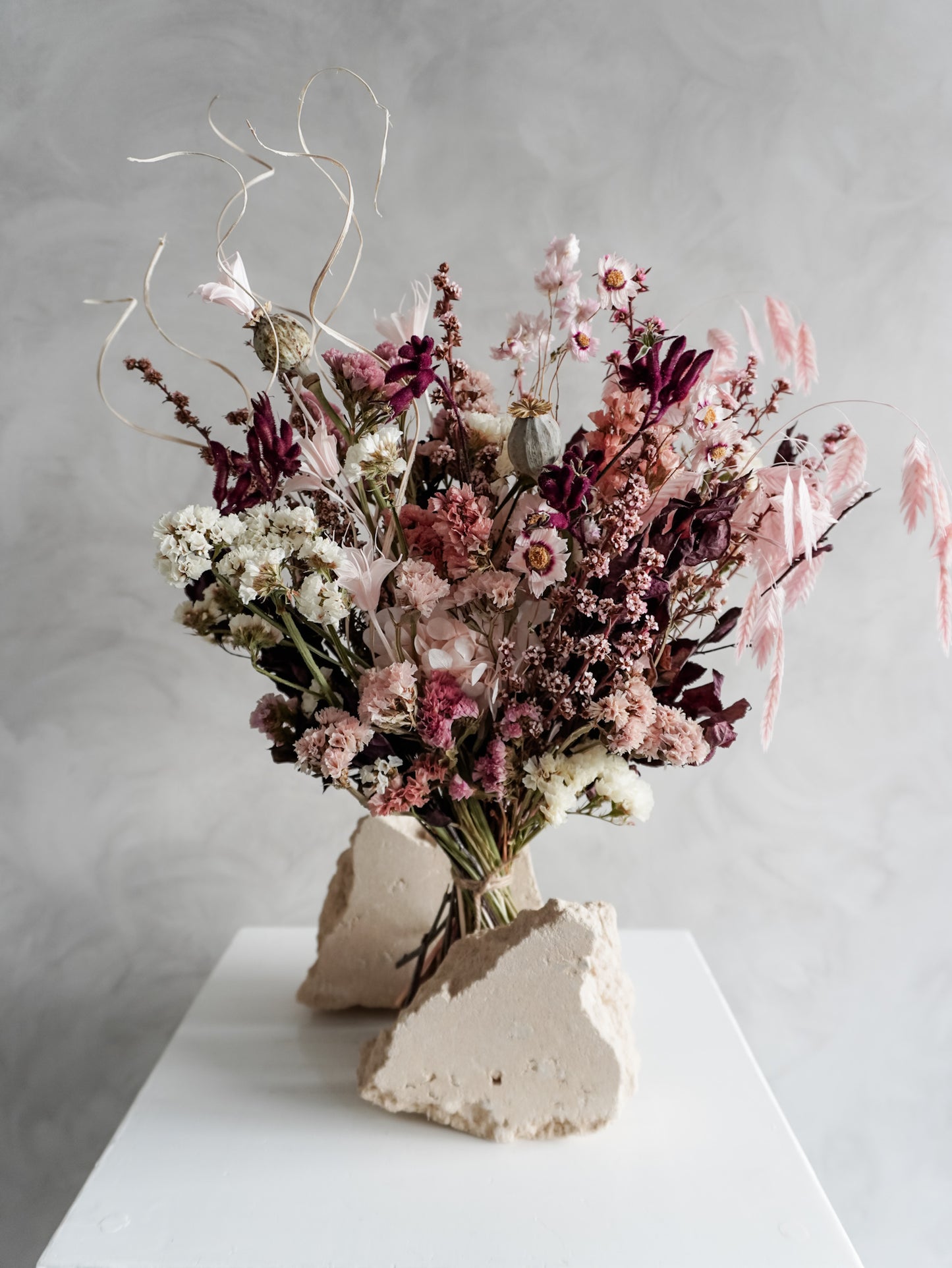 Bouquet of dried flowers in pink and burgundy tones on a white plinth against a grey background.