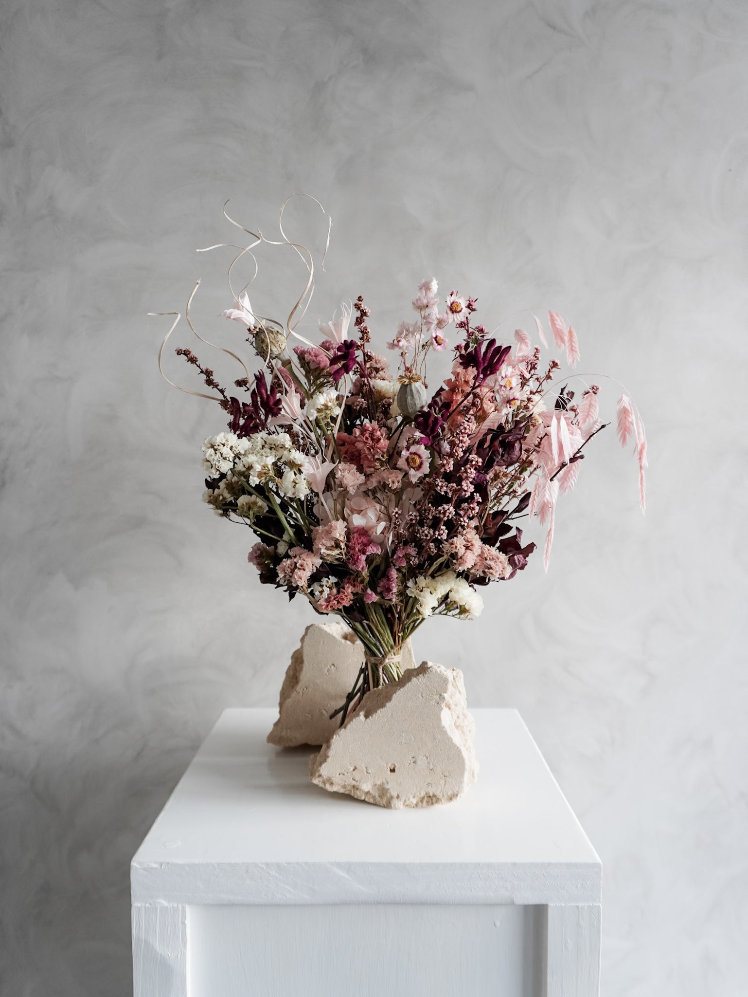 Bouquet of dried flowers in pink and burgundy tones on a white plinth against a grey background.