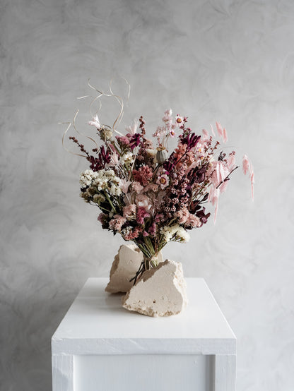 Bouquet of dried flowers in pink and burgundy tones on a white plinth against a grey background.