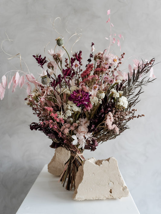 Bouquet of dried flowers in pink and burgundy tones on a white plinth against a grey background.