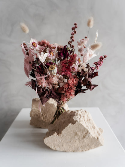 Bouquet of dried flowers in pink and burgundy tones on a white plinth against a grey background.