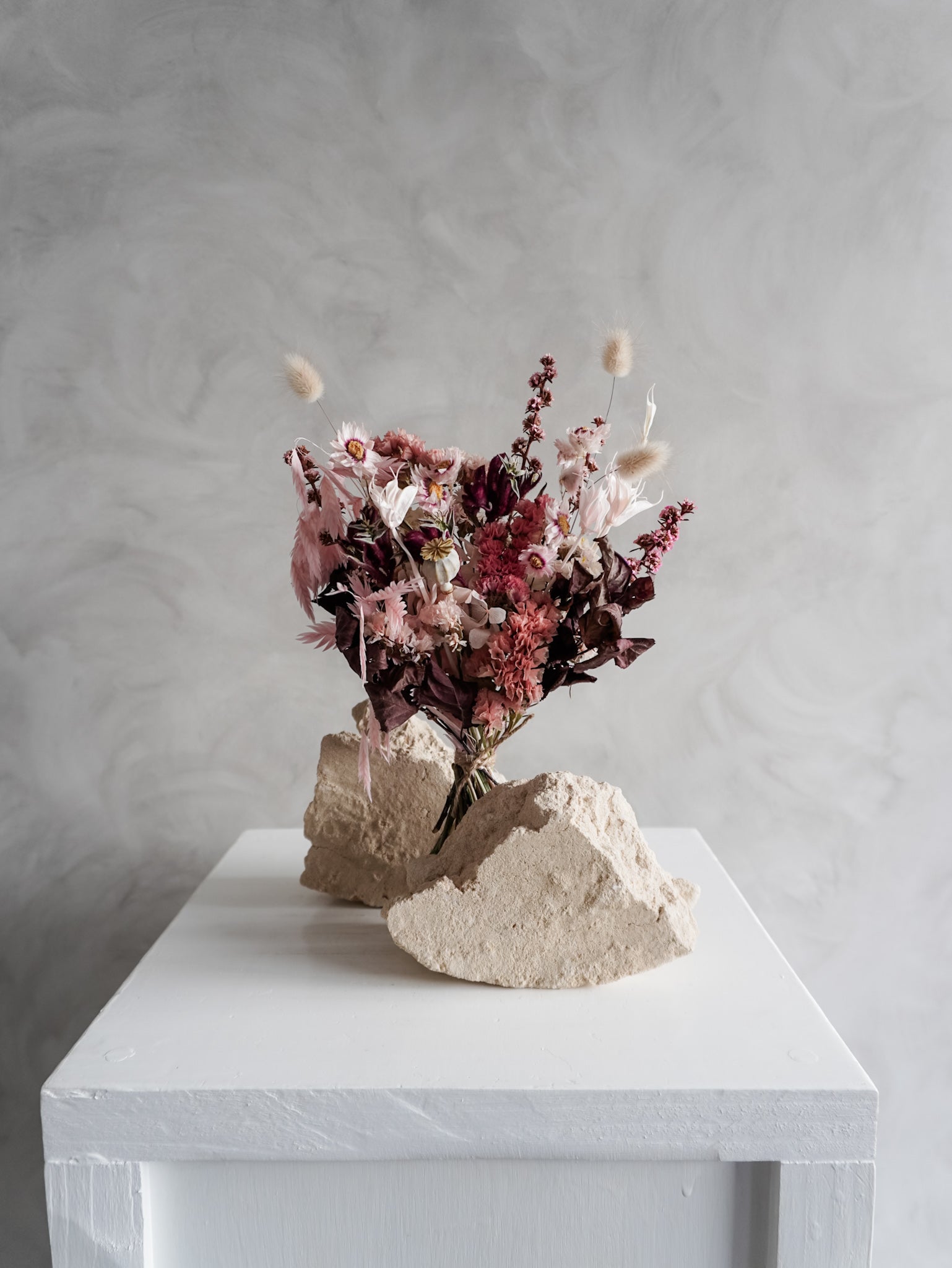 Bouquet of dried flowers in pink and burgundy tones on a white plinth against a grey background.