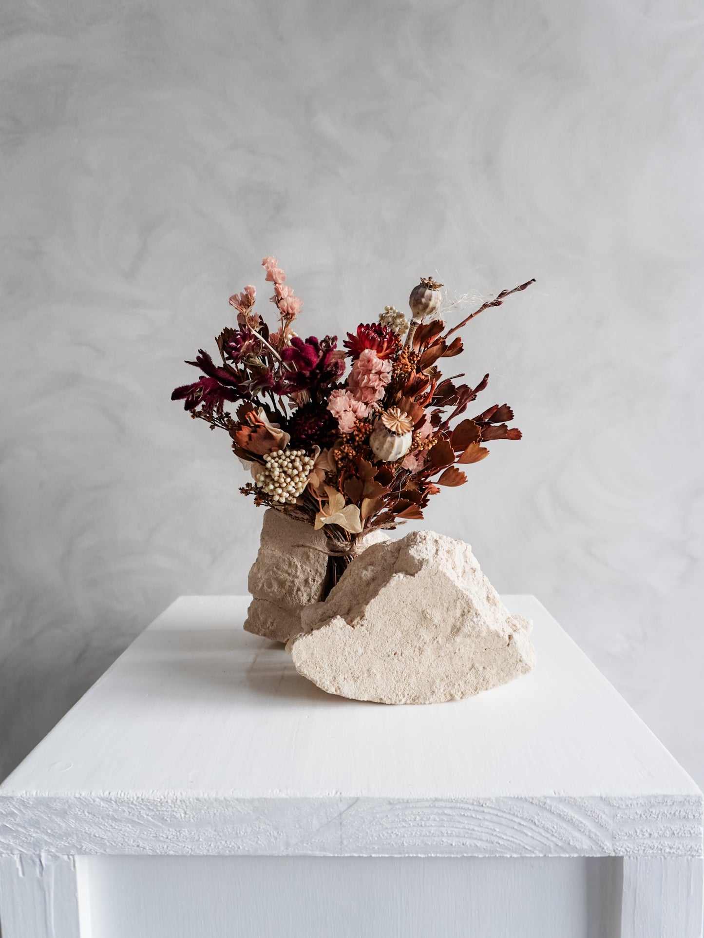 Bouquet of autumnal dried flowers on a white plinth with a grey background - posy size distant view.