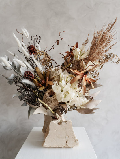 Bouquet of neutral, rustic dried florals on a white plinth against a light gray background.