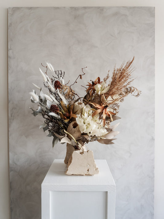 Bouquet of neutral, rustic dried florals on a white plinth against a light gray background.