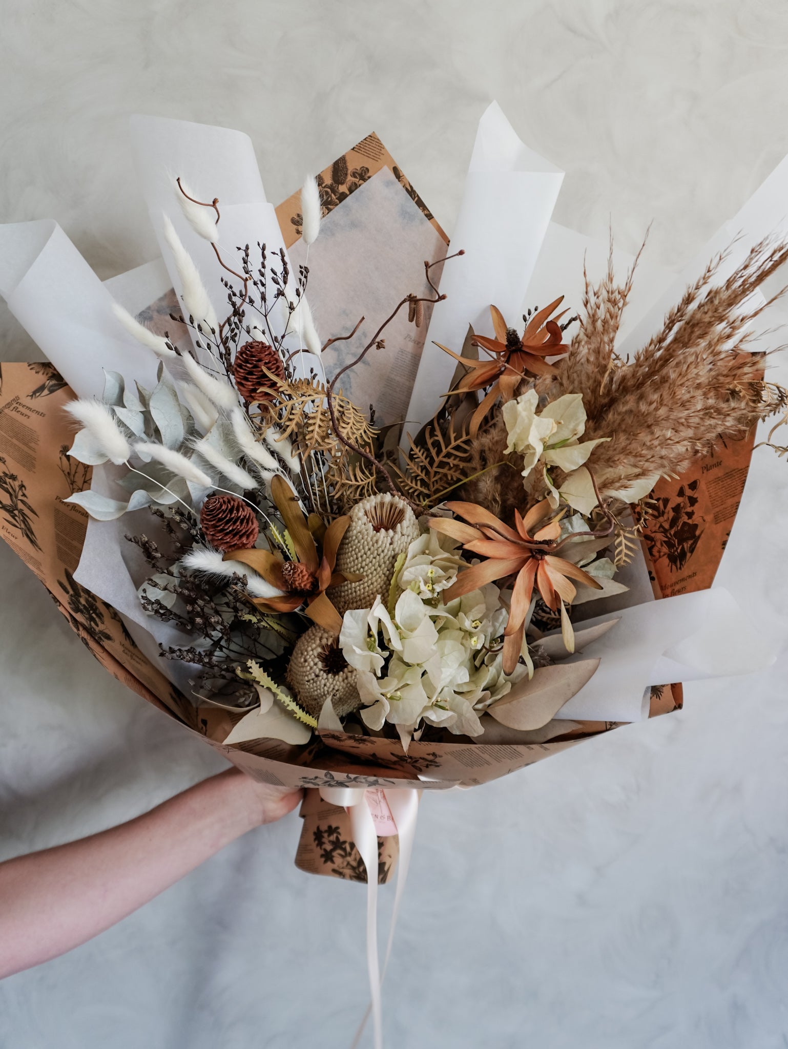 Bouquet of dried flowers wrapped in brown paper held by a hand on a light background.