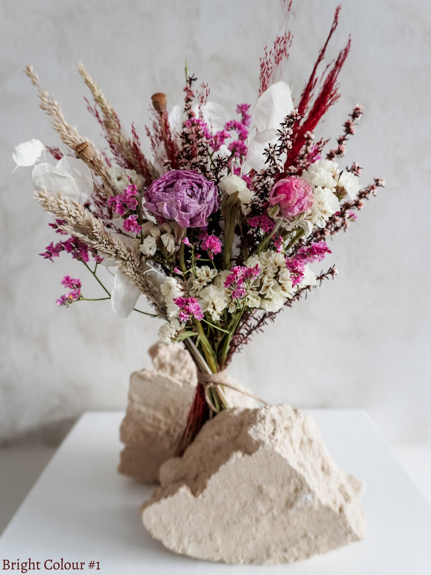 A petite dried flower bouquet featuring pink, purple, white, and red flowers on a white plinth against a light gray background.
