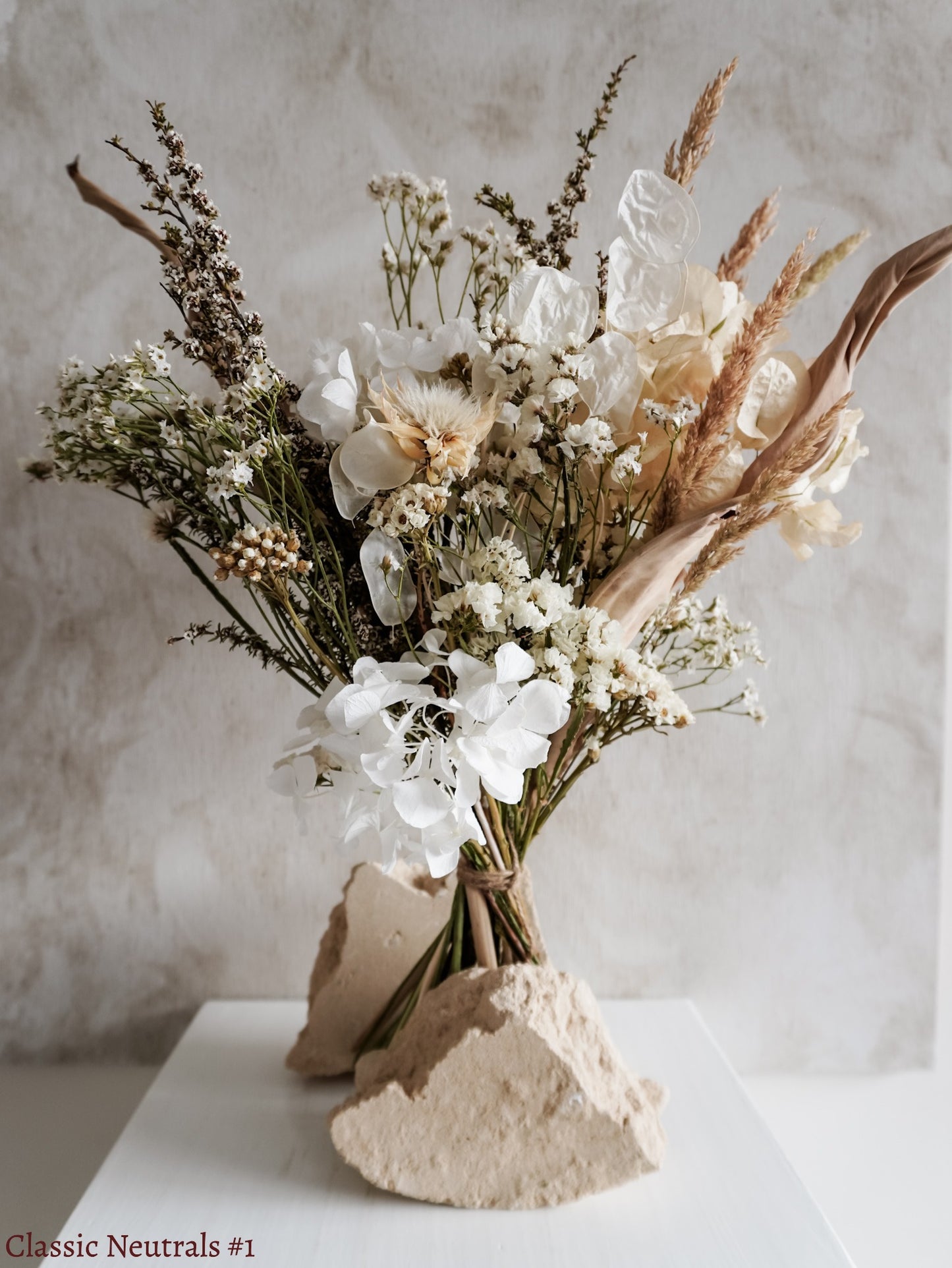A dried flower bouquet featuring white, cream, and neutral flowers against a textured light gray background.