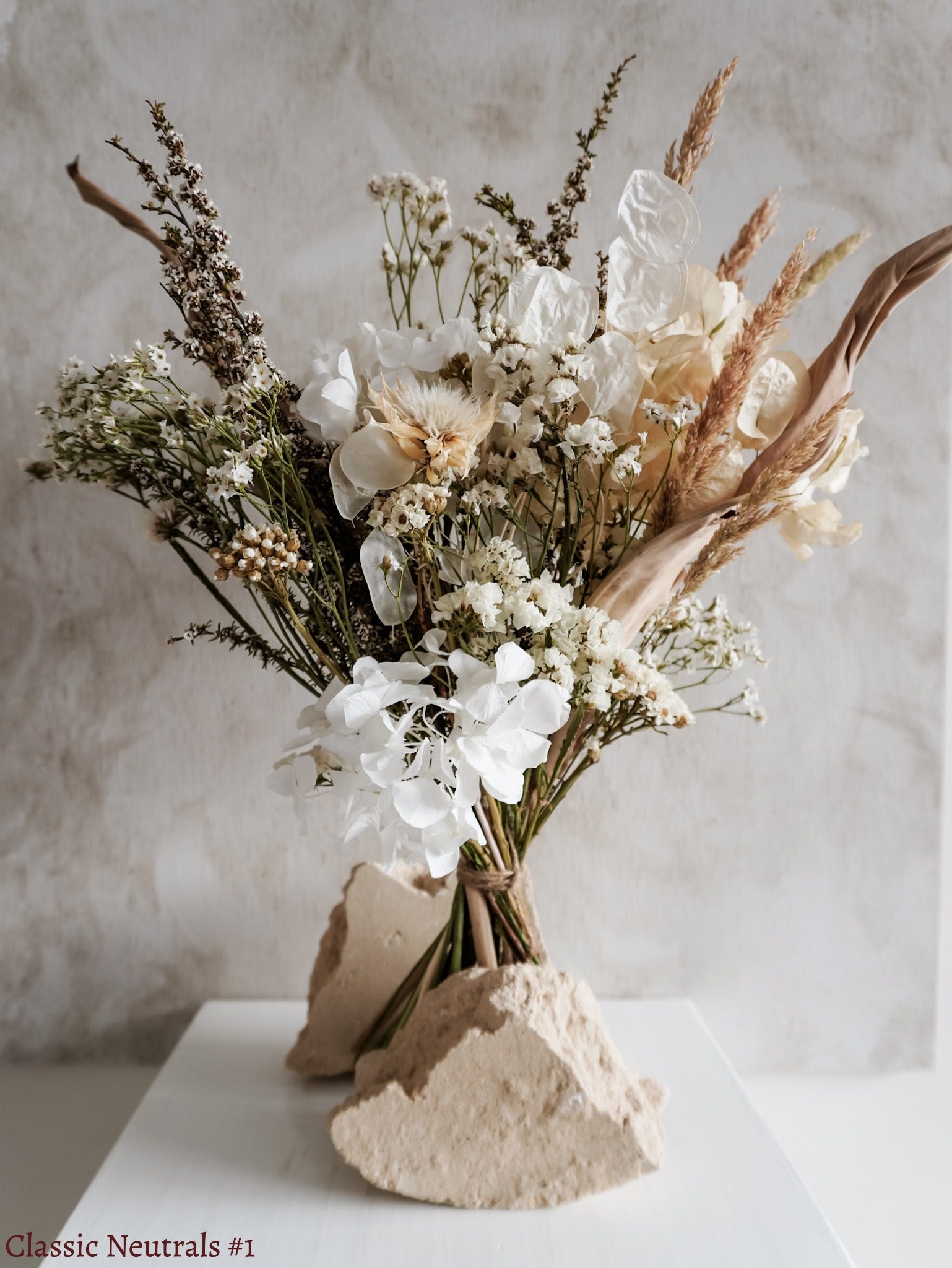 A dried flower bouquet featuring white, cream, and neutral flowers against a textured light gray background.