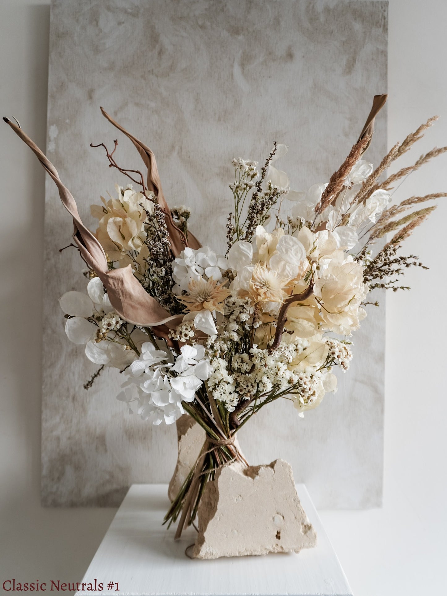 A dried flower bouquet featuring white, cream, and neutral flowers with soft green foliage, displayed on a white plinth against a textured gray background.