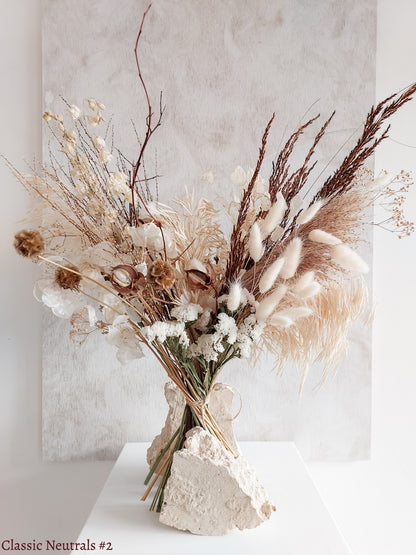 A dried flower bouquet featuring white, cream, and earthy tone flowers, displayed on a white plinth against a textured gray background.