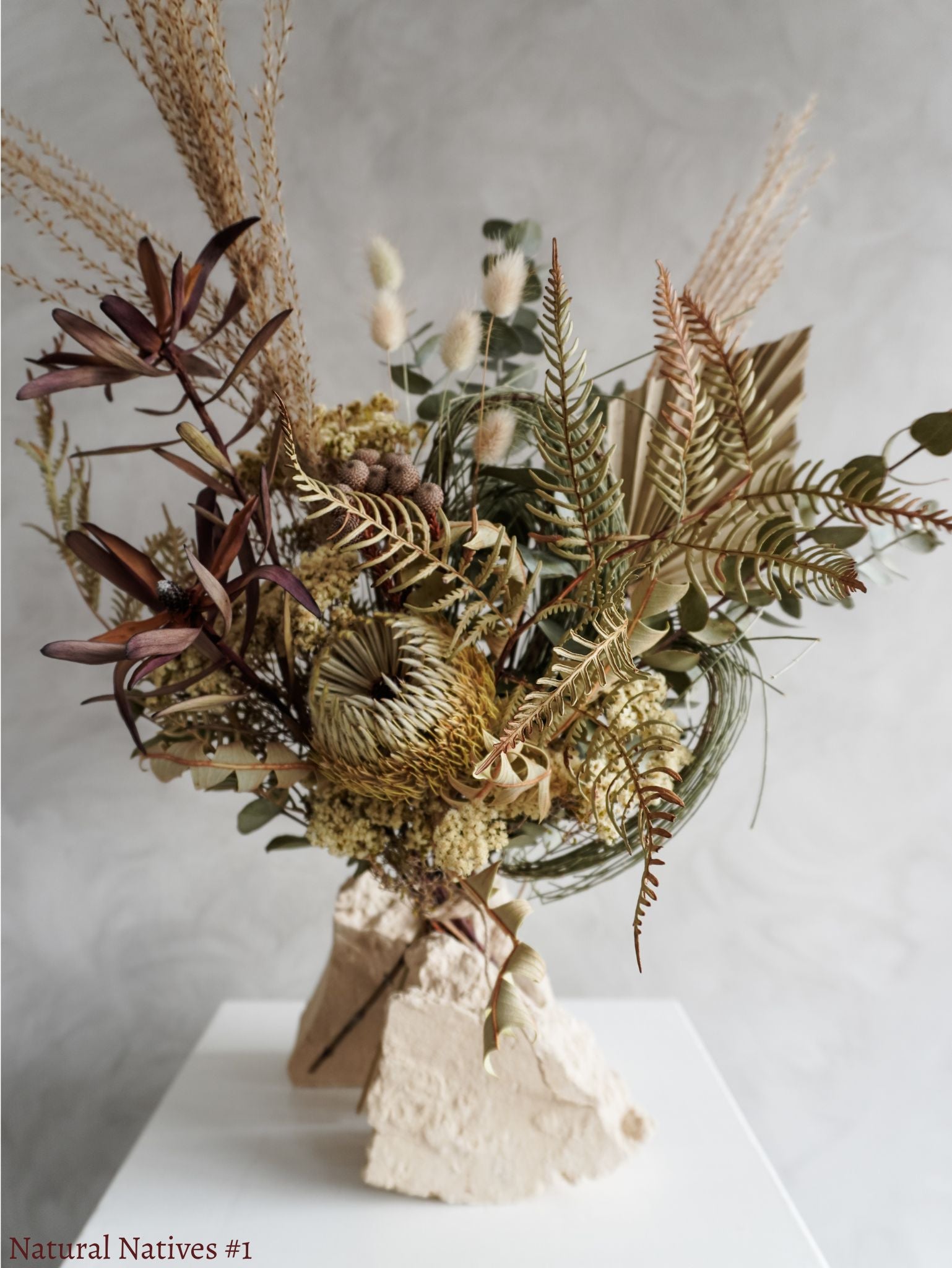 A dried flower bouquet featuring textural native flowers in neutral tones against a textured light gray background.