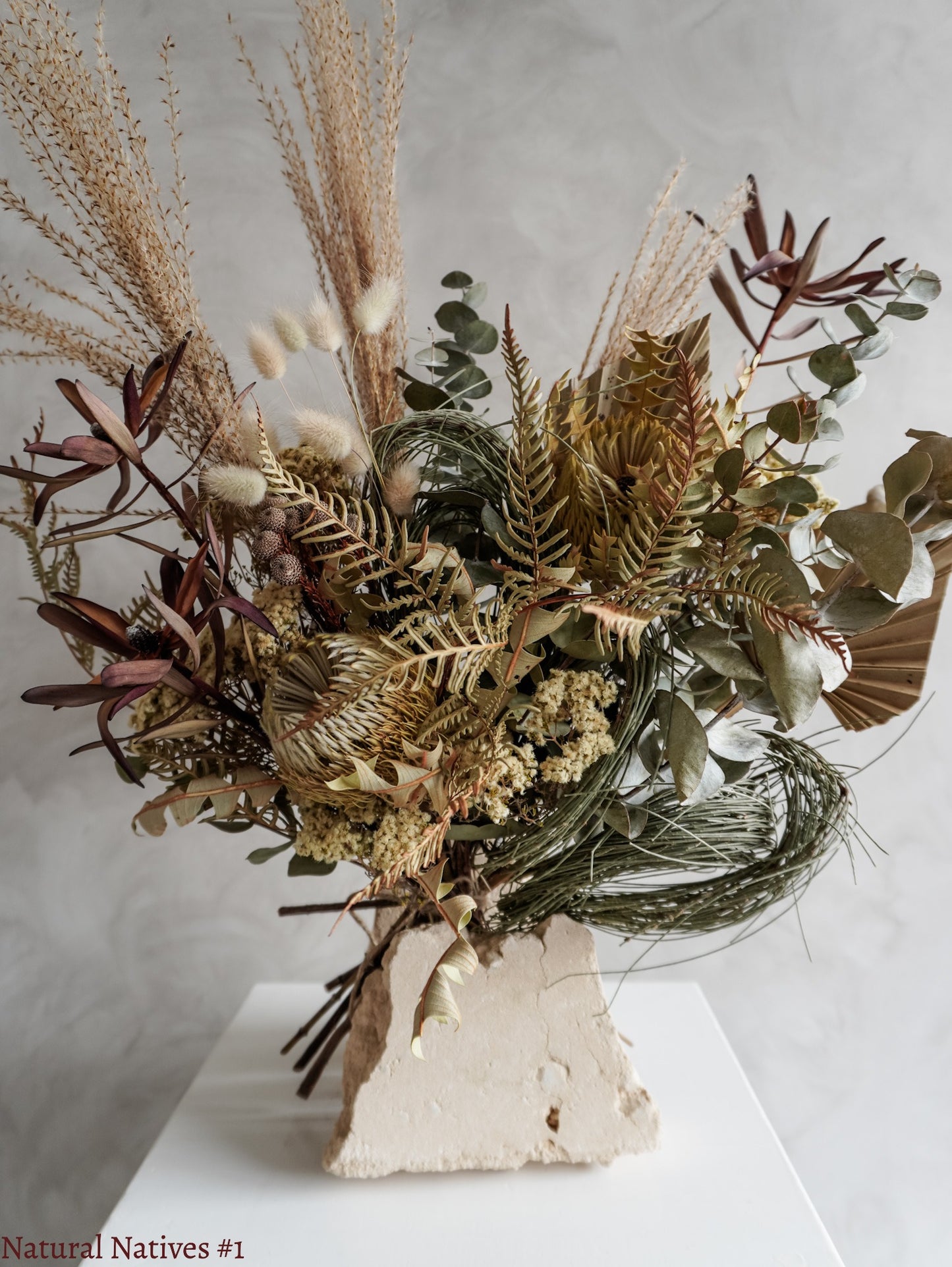 A dried flower bouquet featuring textural native flowers in neutral tones, displayed on a white plinth against a textured gray background.