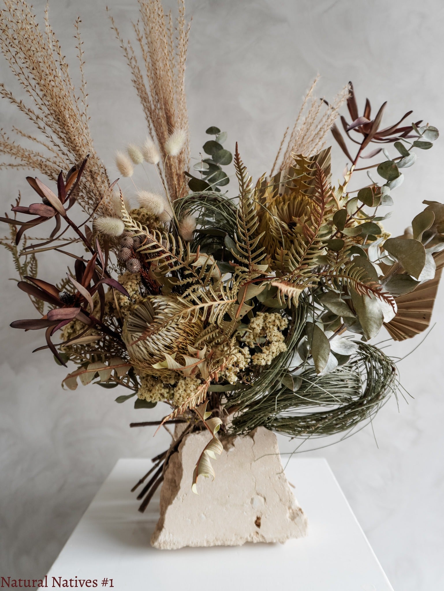 A dried flower bouquet featuring textural native flowers in neutral tones, displayed on a white plinth against a textured gray background.