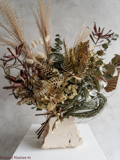 A dried flower bouquet featuring textural native flowers in neutral tones, displayed on a white plinth against a textured gray background.