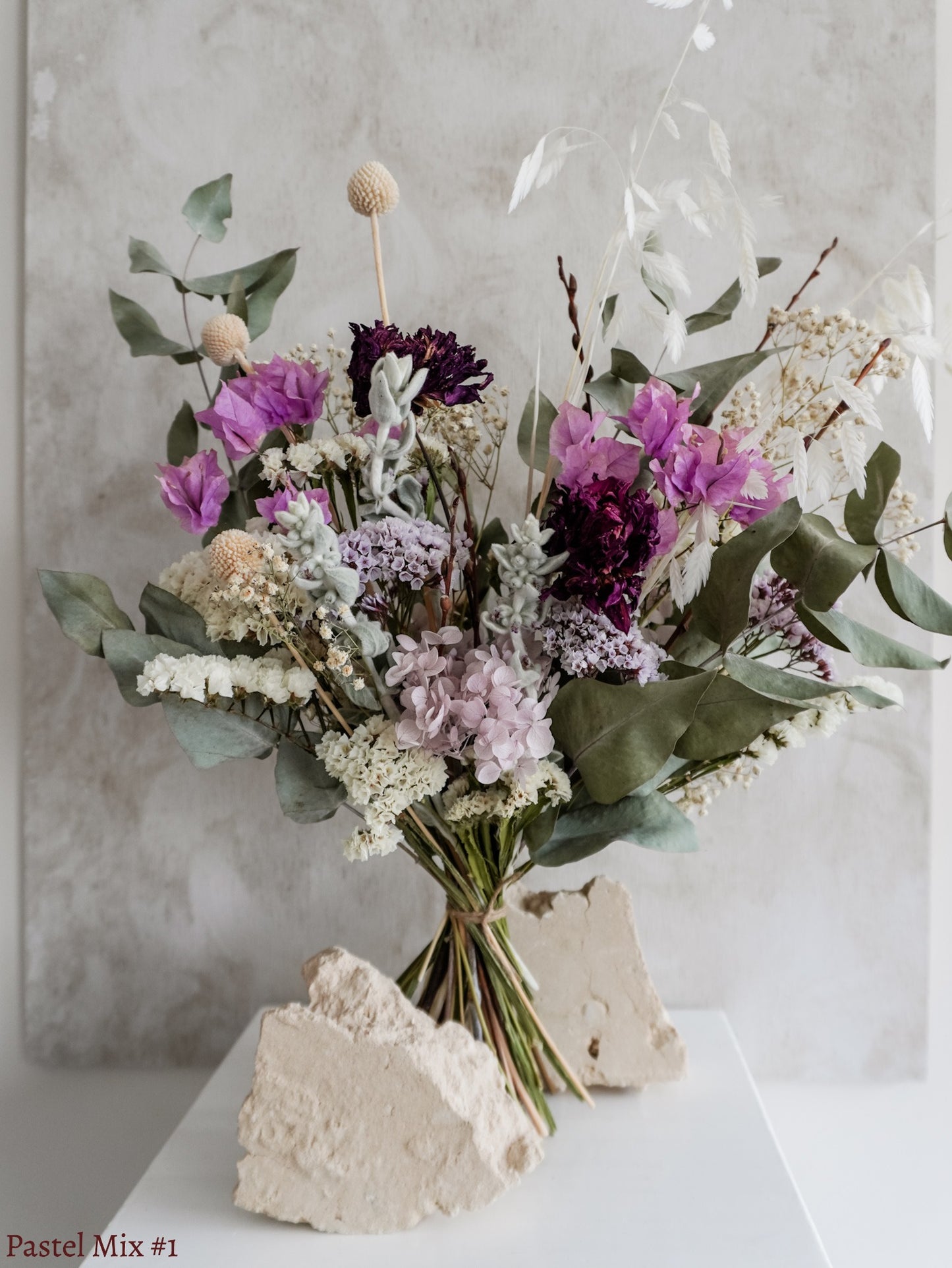A dried flower bouquet featuring mauve, soft green, and white flowers against a textured light gray background.