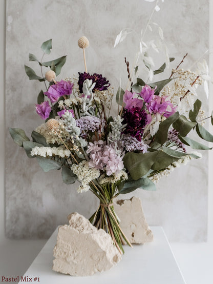 A dried flower bouquet featuring mauve, soft green, and white flowers against a textured light gray background.