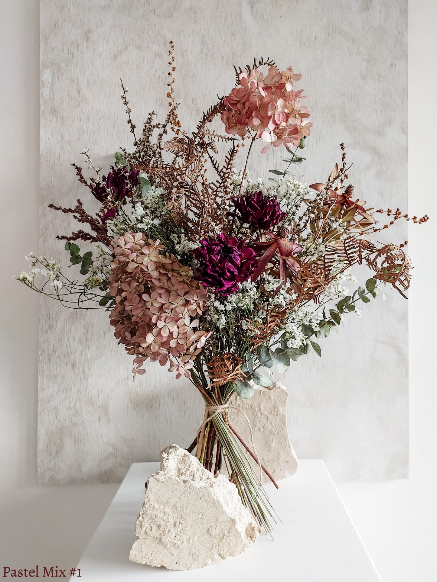 A dried flower bouquet featuring soft pink, white, purple flowers with forest green foliage, displayed on a white plinth against a textured gray background.