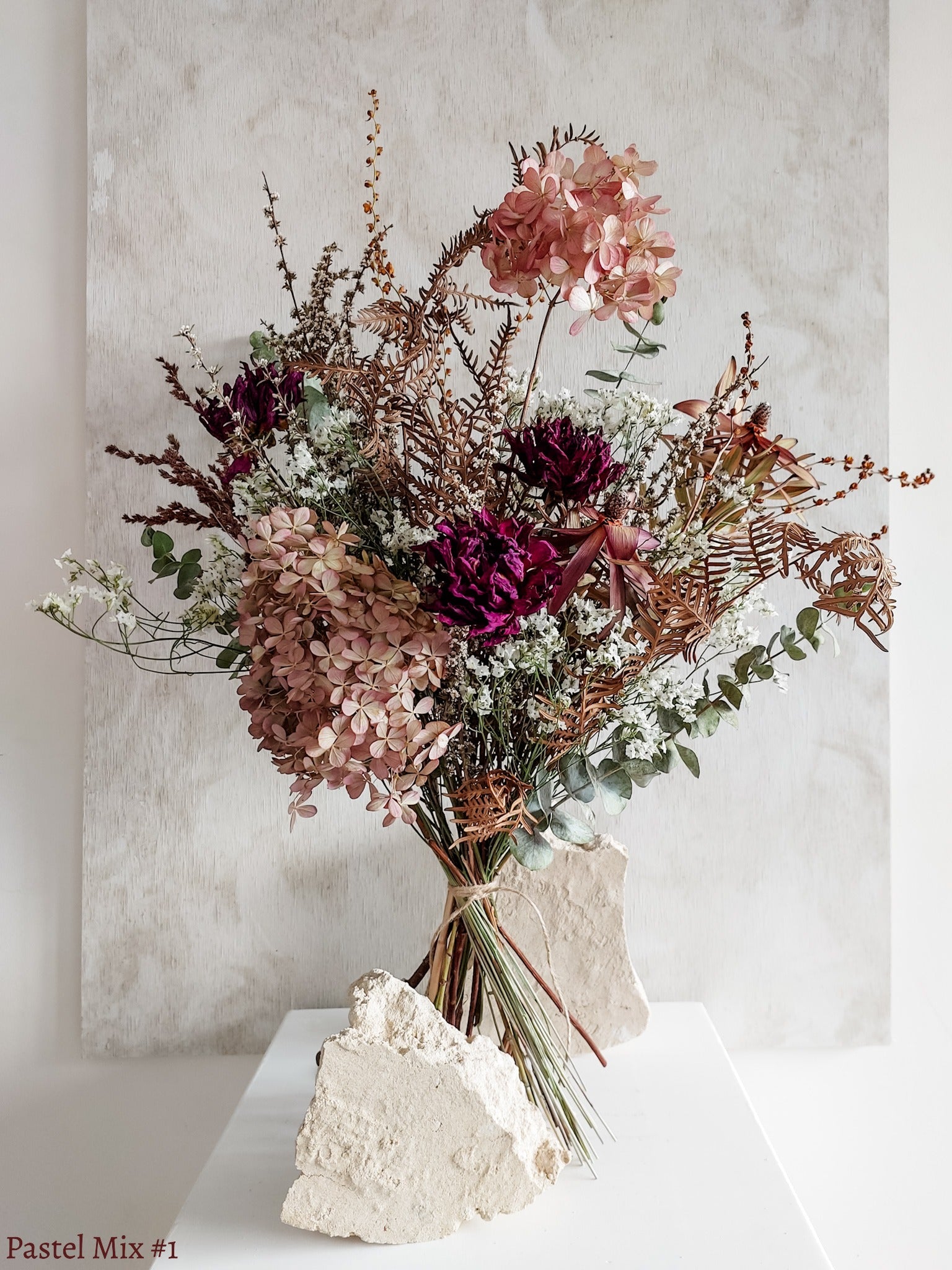A dried flower bouquet featuring soft pink, white, purple flowers with forest green foliage, displayed on a white plinth against a textured gray background.