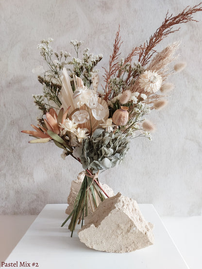 A dried flower bouquet featuring white, earthy, and soft blue flowers against a textured light gray background.